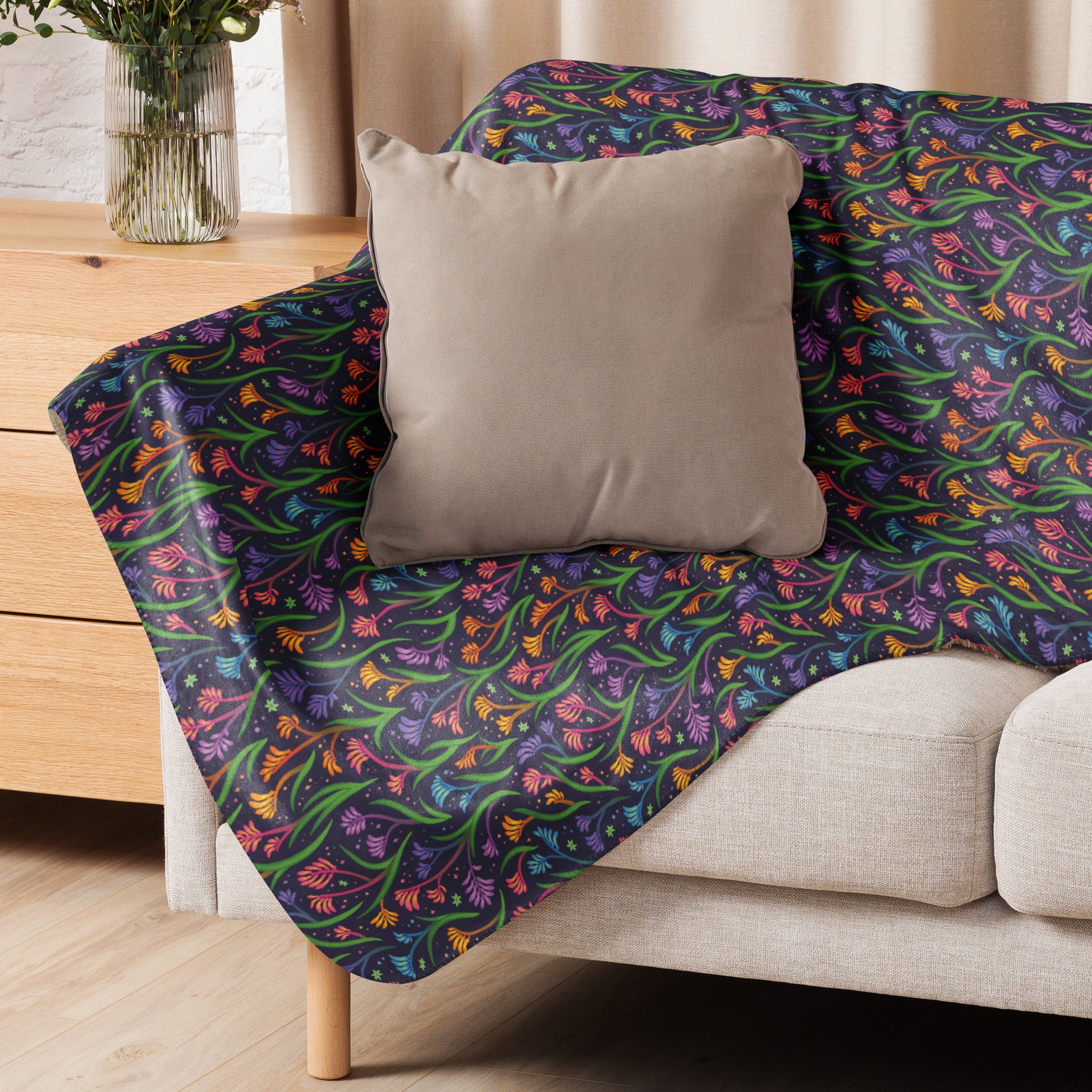 A rainbow kangaroo paw flower pattern blanket sitting on a beige sofa with a beige pillow on top.