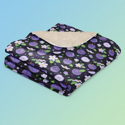 A folded purple apples & blossoms pattern blanket on a blue and green gradient background.