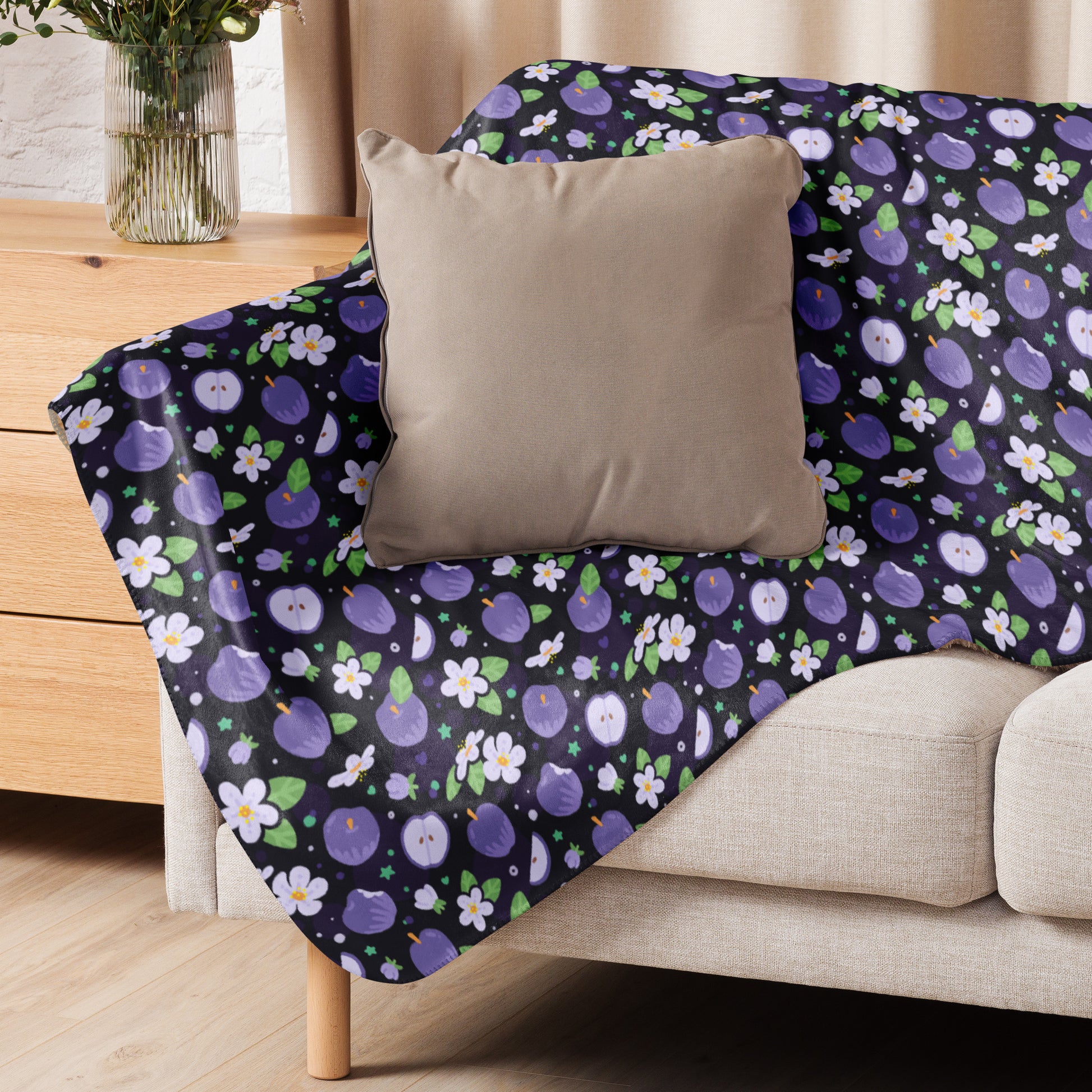 A purple apples & blossoms pattern blanket sitting on a beige sofa with a beige pillow on top.