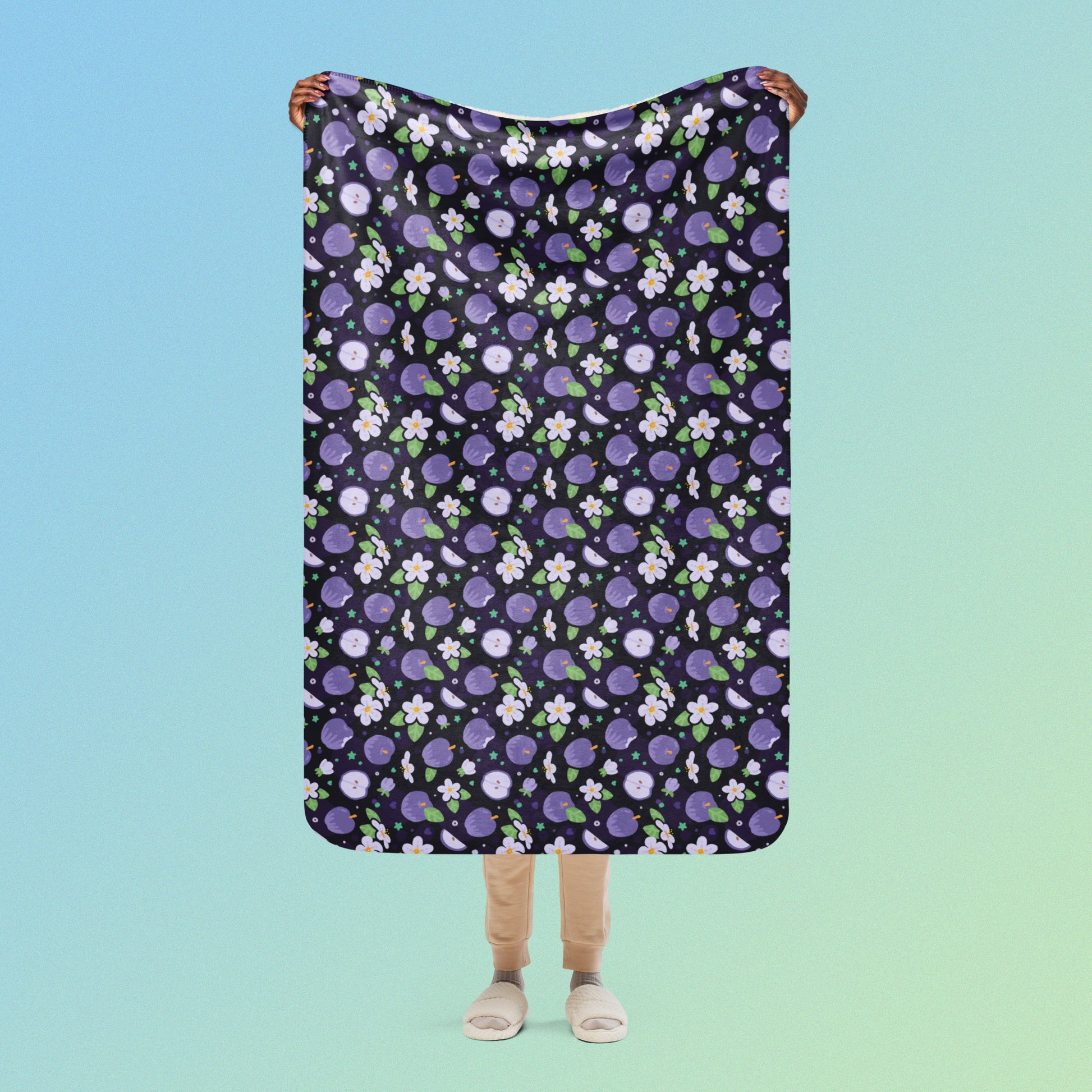 Person holding a purple apples & blossoms pattern blanket above their head on a blue and green gradient background.