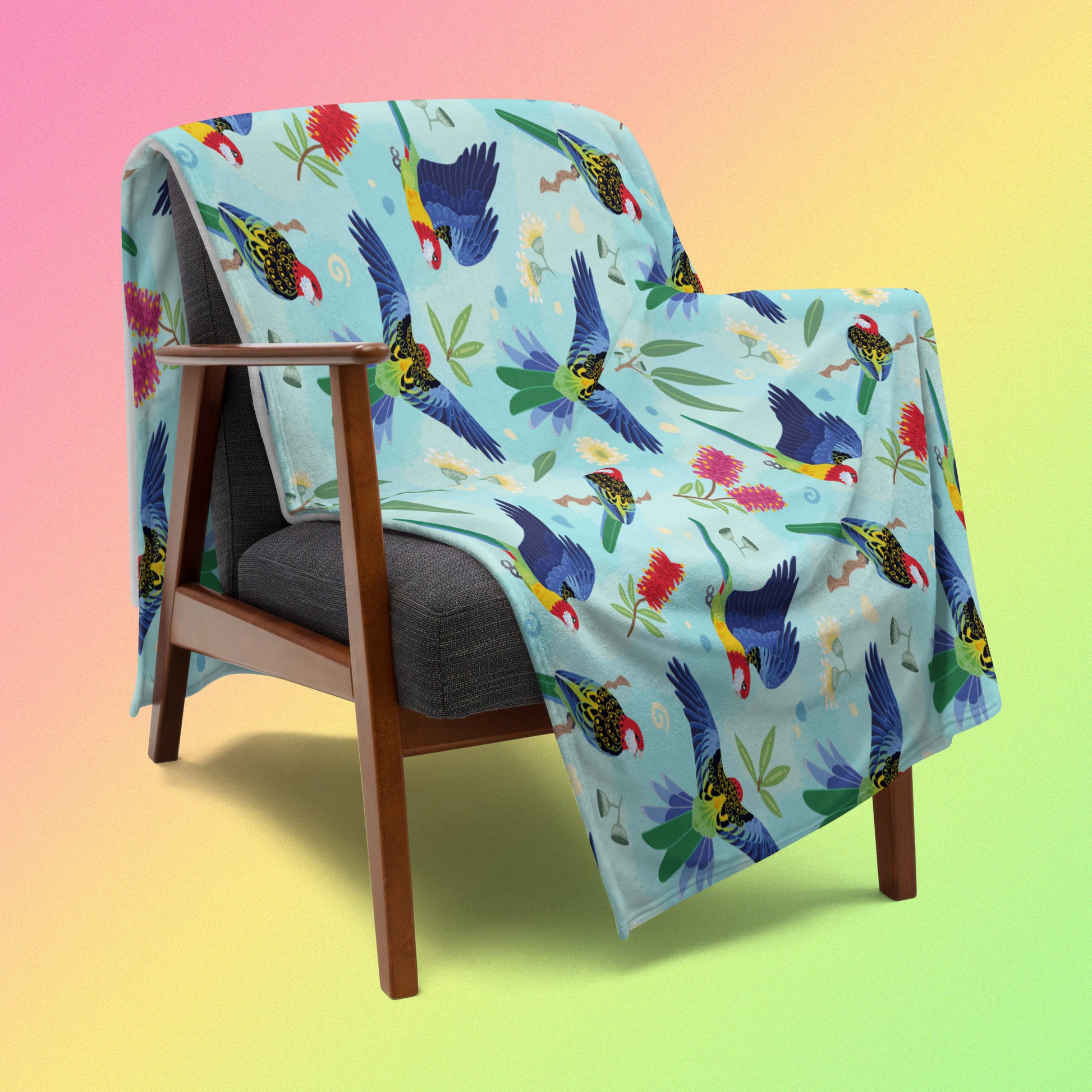 A blue, pink and green eastern rosella & native Aussie plant pattern blanket draped over a wooden chair on a pink, yellow and green gradient background.