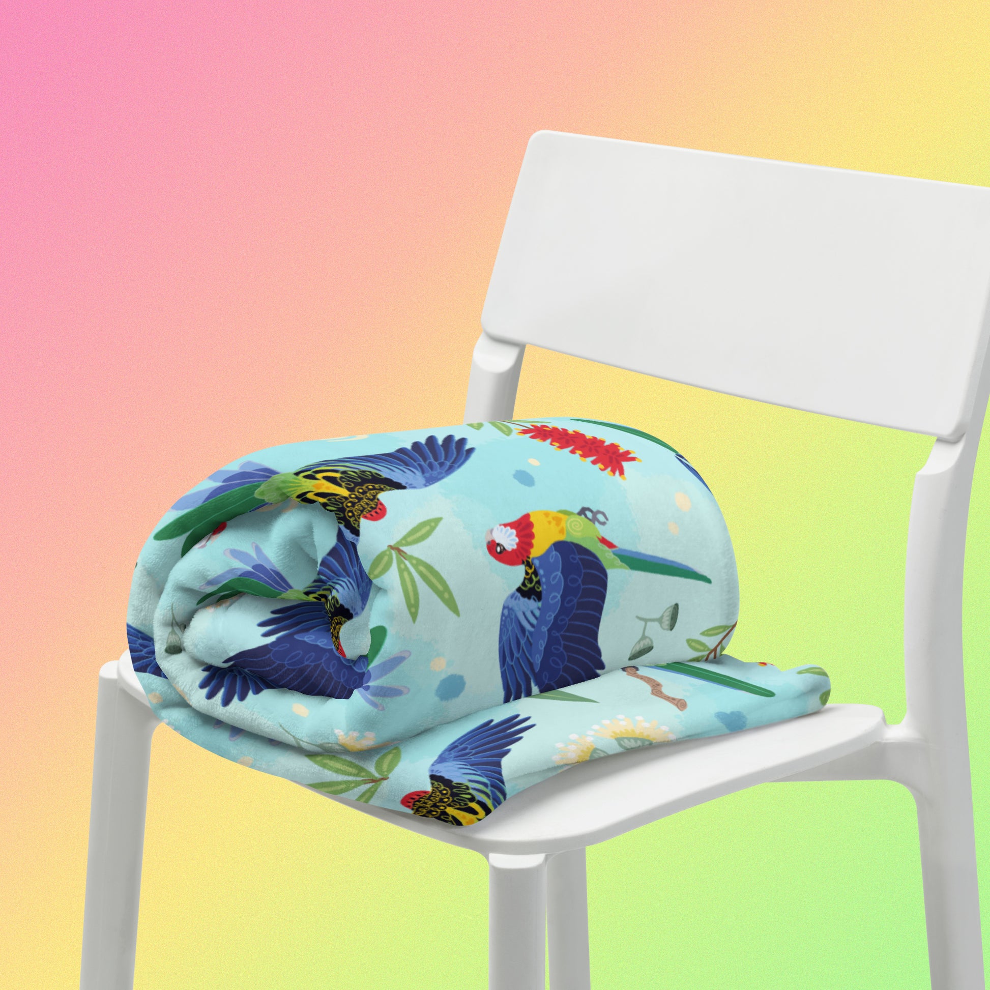 A rolled up blue, pink and green eastern rosella & native Aussie plant pattern blanket sitting on a white chair on a pink, yellow and green gradient background.