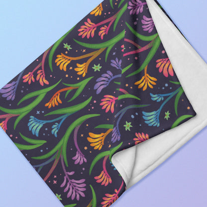 Close up detail of a folded rainbow kangaroo paw flower pattern blanket on a blue and purple gradient background.