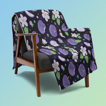 A purple apples & blossoms pattern blanket draped over a wooden chair on a blue and green gradient background.