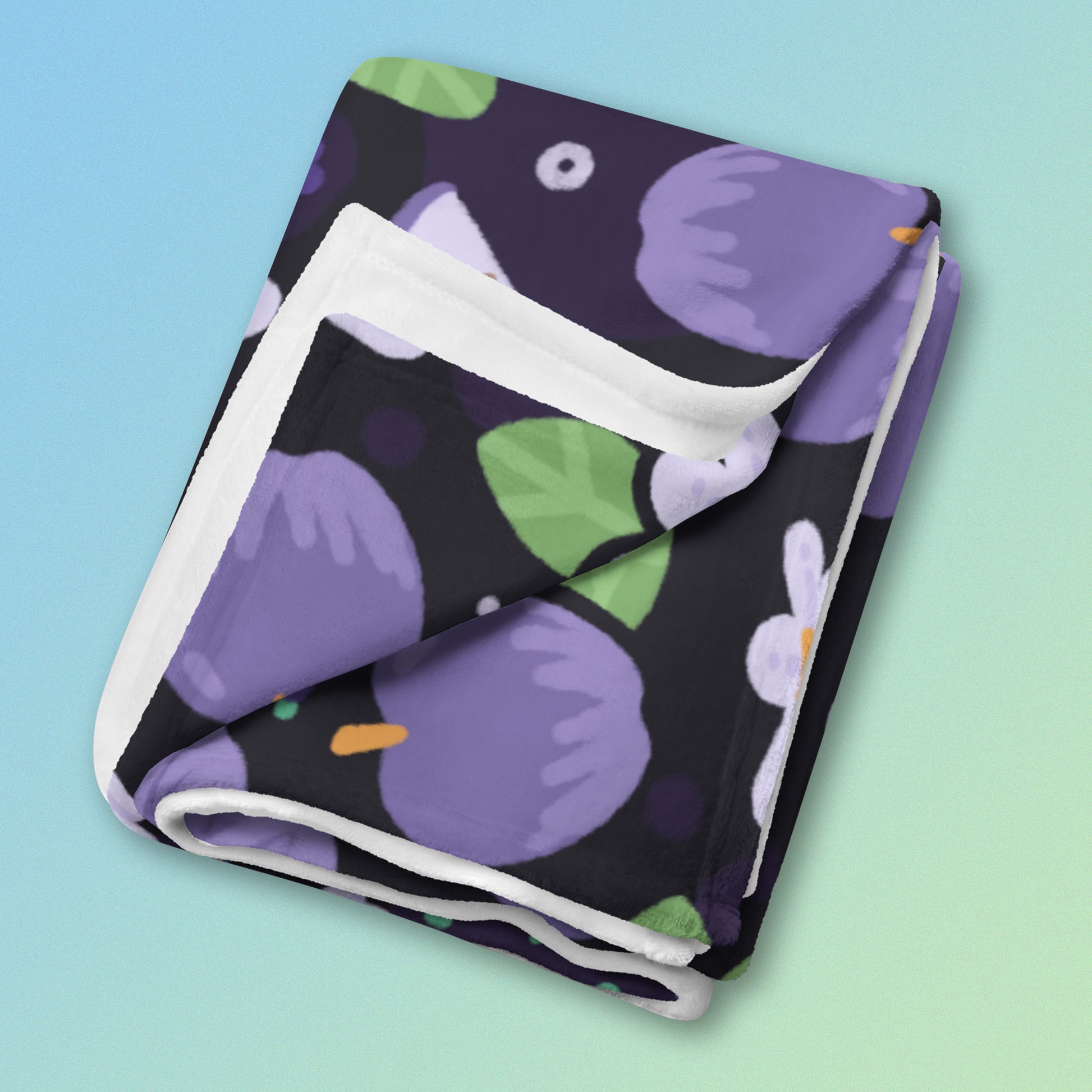 A folded purple apples & blossoms pattern blanket on a blue and green gradient background.