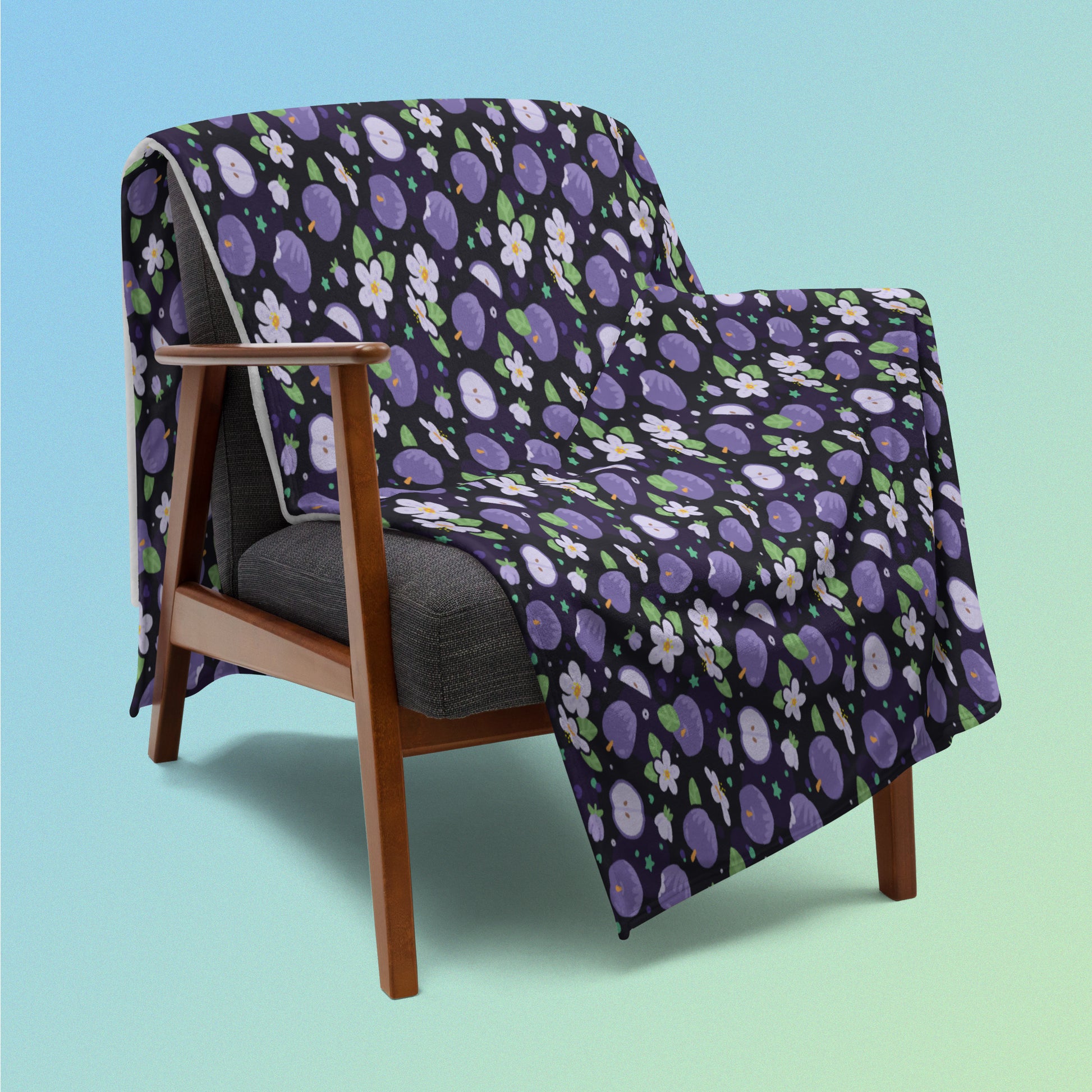 A purple apples & blossoms pattern blanket draped over a wooden chair on a blue and green gradient background.