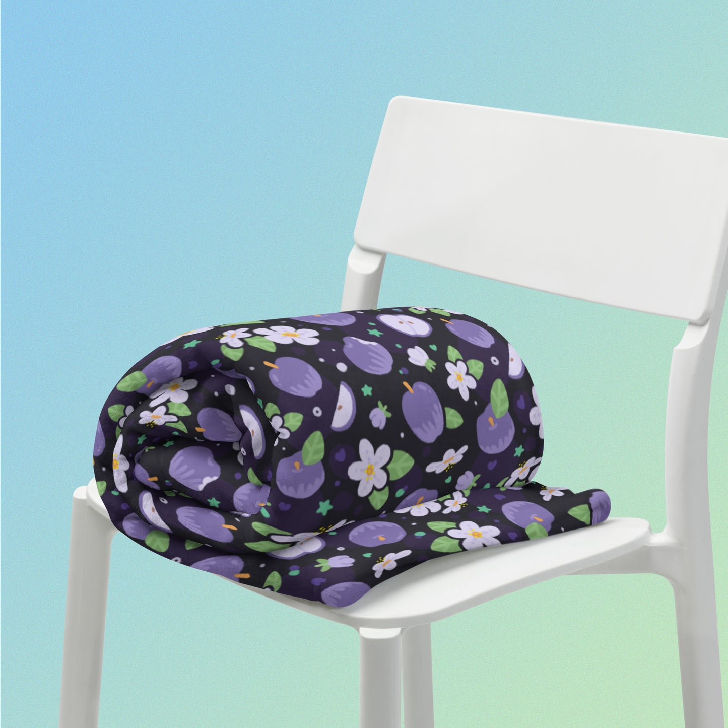 A rolled up purple apples & blossoms pattern blanket sitting on a white chair on a blue and green gradient background.