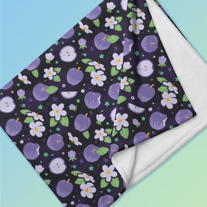 Close up detail of a folded purple apples & blossoms pattern blanket on a blue and green gradient background.