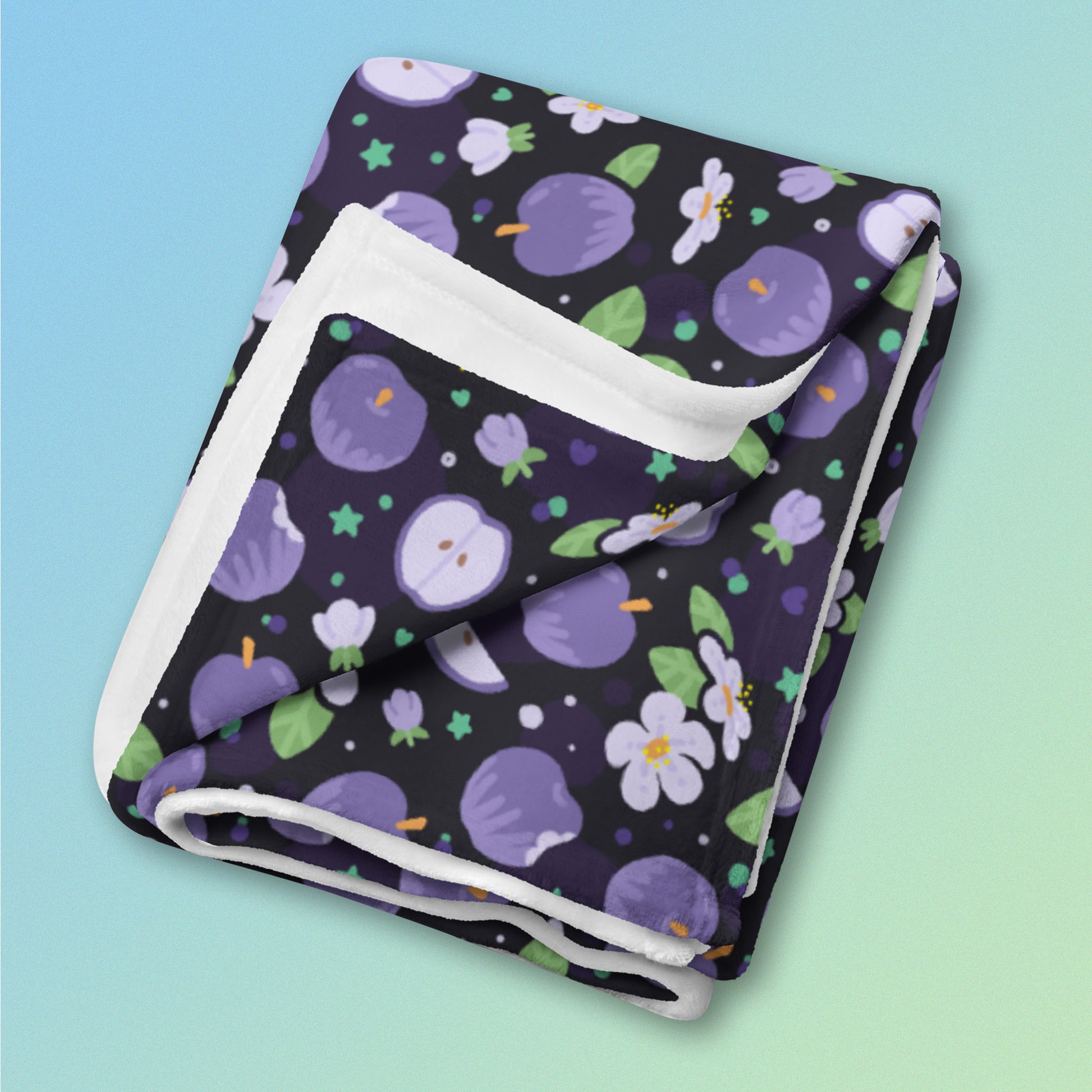 A folded purple apples & blossoms pattern blanket on a blue and green gradient background.