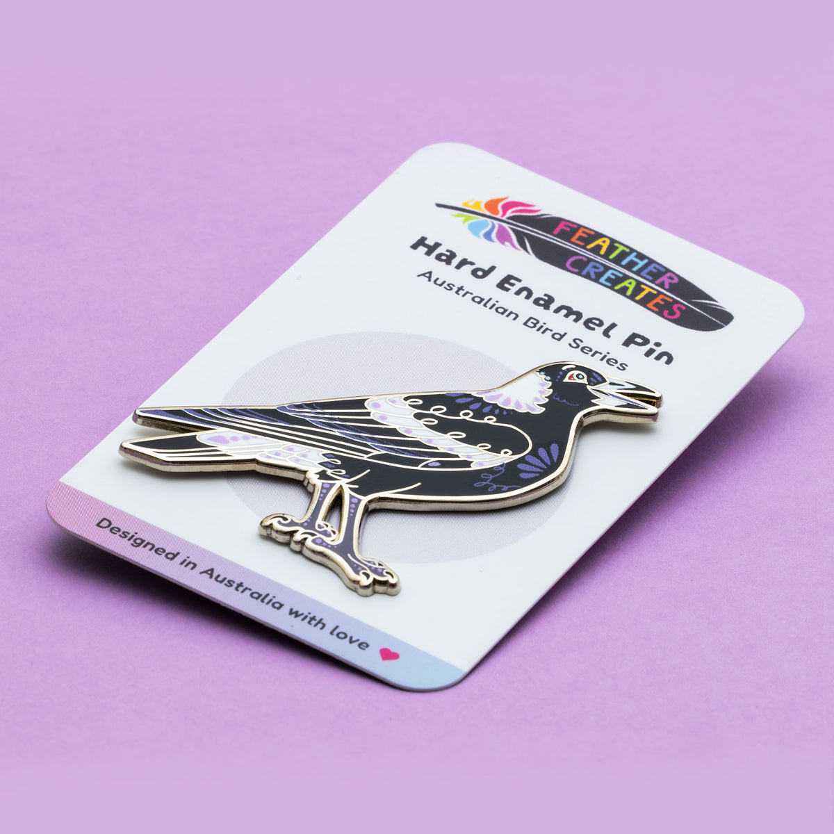 A nickel-plated Australian magpie hard enamel pin on a 'Feather Creates' branded backing card sitting on an angle on a purple background.