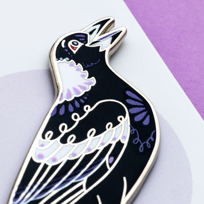 Close up on the face of a nickel-plated Australian magpie hard enamel pin on a 'Feather Creates' branded backing card sitting on an angle on a purple background.
