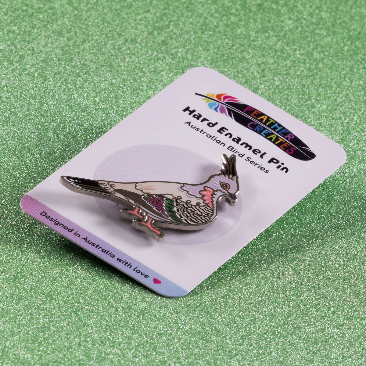 A nickel-plated crested pigeon hard enamel pin sitting on a 'Feather Creates' branded backing card on green glitter background.