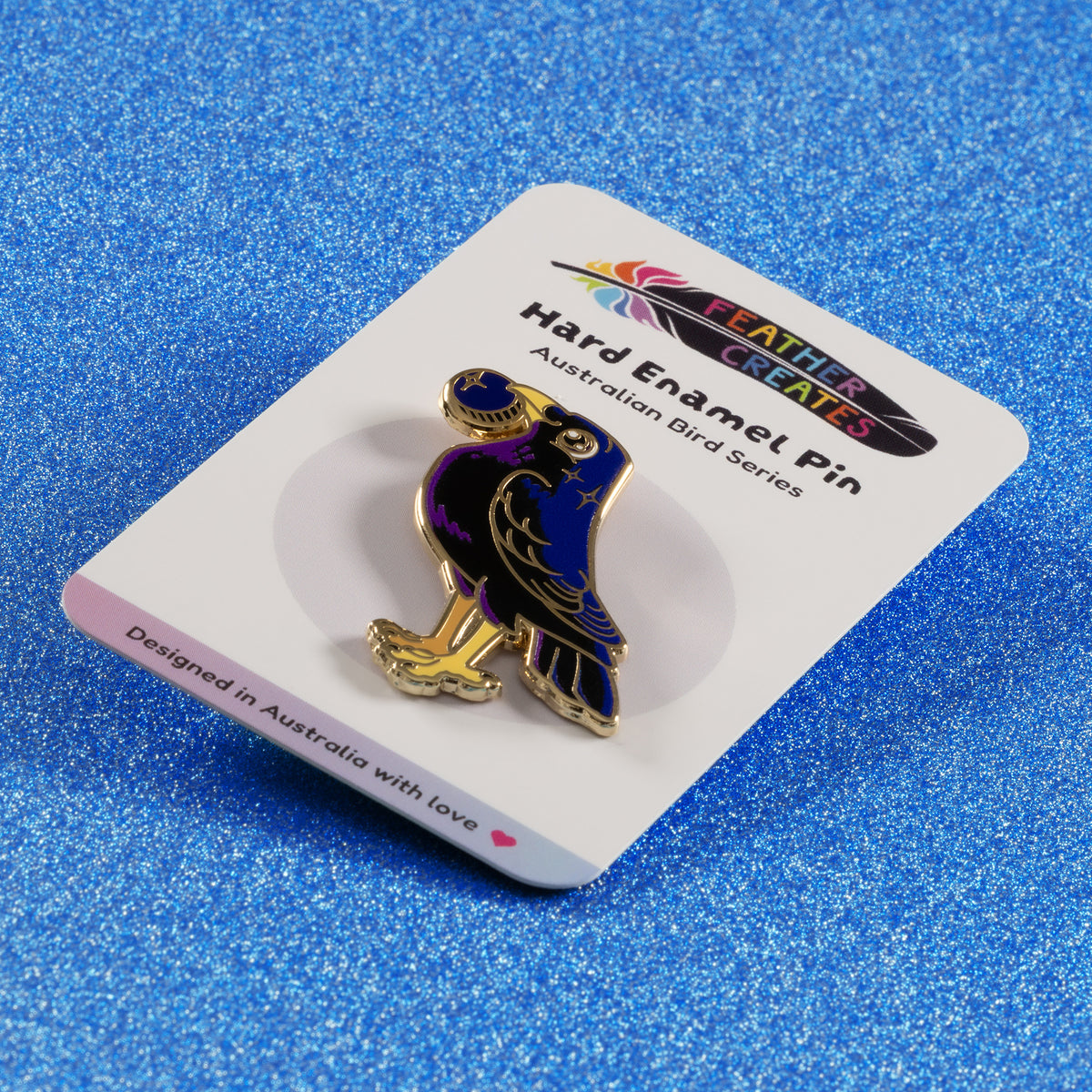 A gold-plated satin bowerbird hard enamel pin sitting on a 'Feather Creates' branded backing card on a blue glitter background.