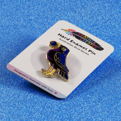 A gold-plated satin bowerbird hard enamel pin sitting on a 'Feather Creates' branded backing card on a blue glitter background.