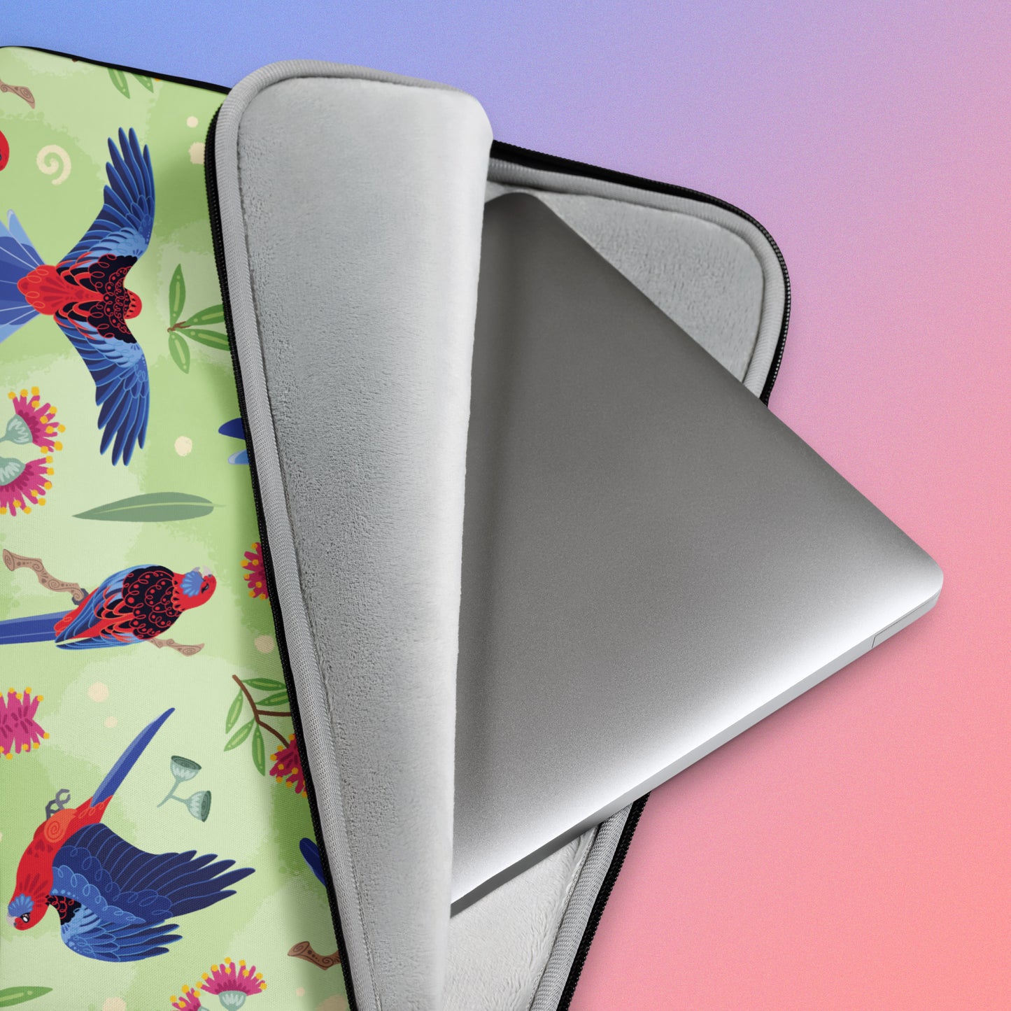 Close up detail of a laptop sitting in a laptop sleeve with a colourful green, red, blue and pink crimson rosella pattern on a pink and blue gradient background.