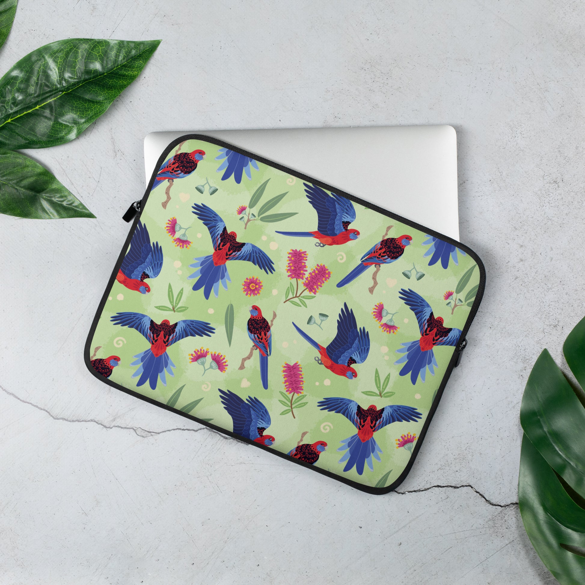 Laptop sleeve with a colourful green, red, blue and pink crimson rosella pattern sitting on a marble table surrounded by leaves.