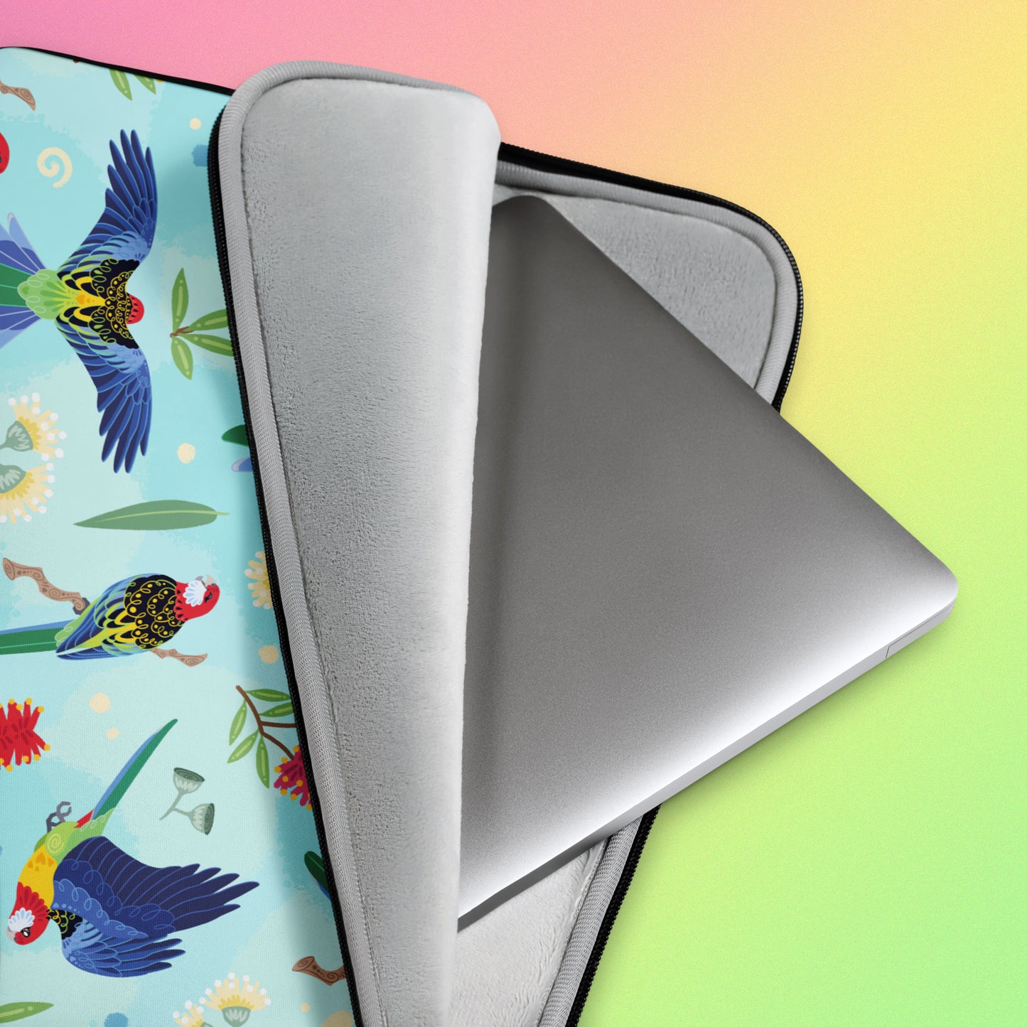 Close up detail of a laptop sitting in a laptop sleeve with a colourful rainbow, blue, green, red and pink Eastern Rosella pattern on a pink, yellow and green gradient background.