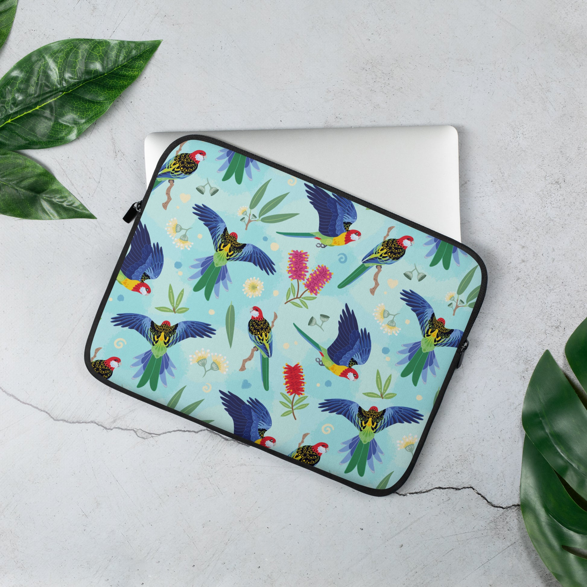 Laptop sleeve with a colourful rainbow, blue, green, red and pink Eastern Rosella pattern sitting on a marble table surrounded by leaves.