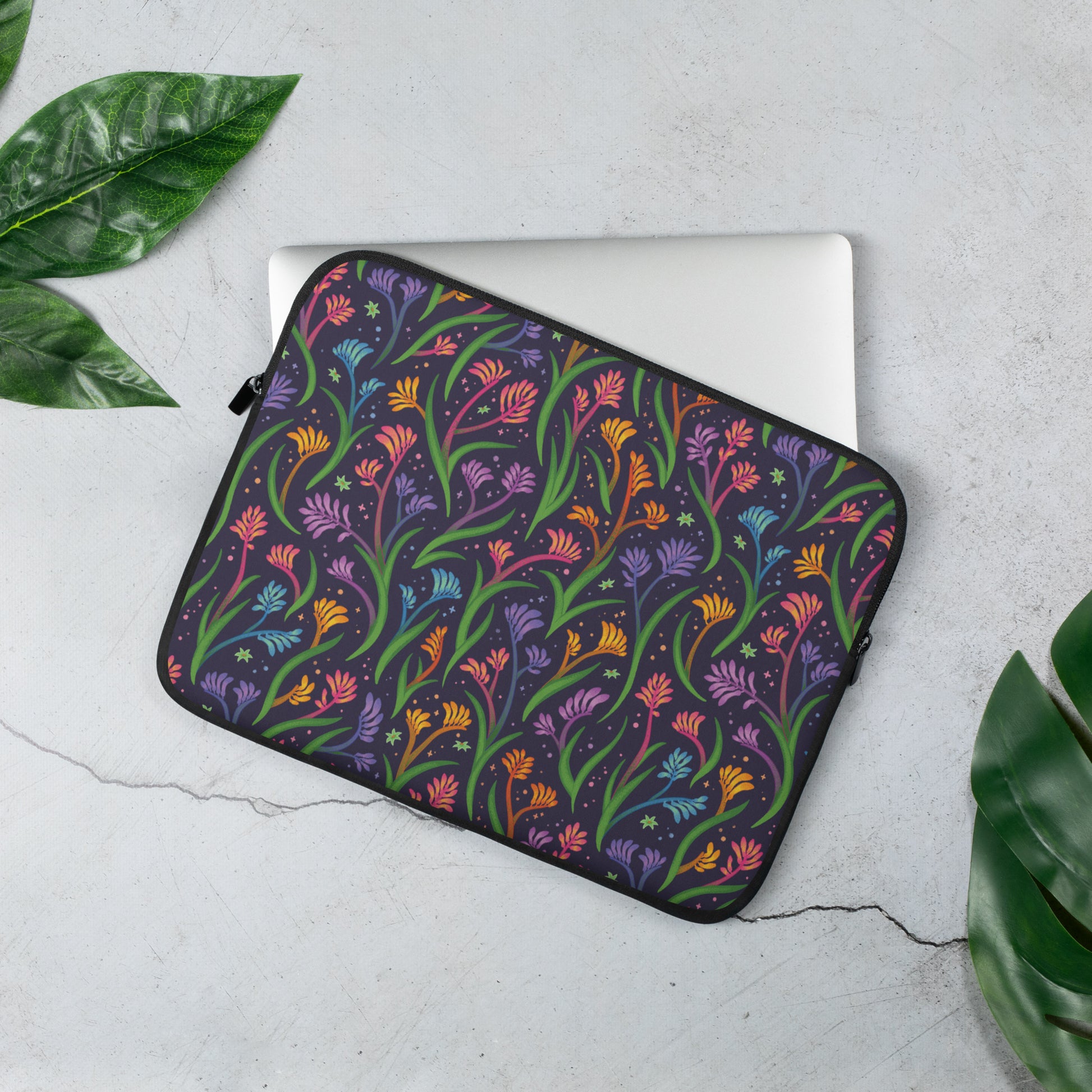 Laptop sleeve with a colourful rainbow kangaroo paw flower pattern sitting on a marble table surrounded by leaves.