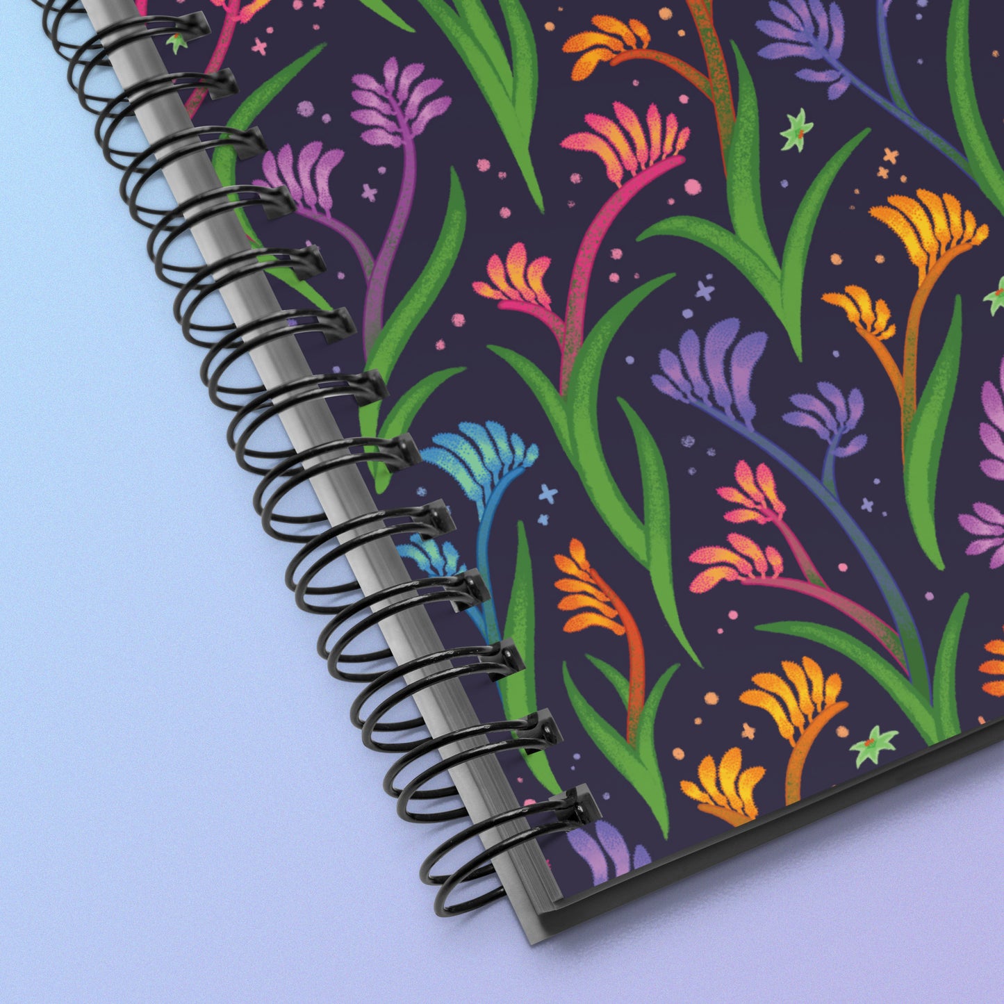 Close up of a spiral notebook with a colourful rainbow kangaroo paw flower pattern on a pink and purple gradient background.