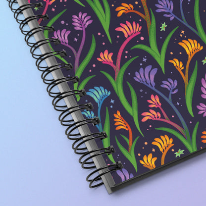 Close up of a spiral notebook with a colourful rainbow kangaroo paw flower pattern on a pink and purple gradient background.