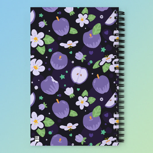 Spiral notebook with a colourful purple apples and apple blossoms pattern on a blue and green gradient background.