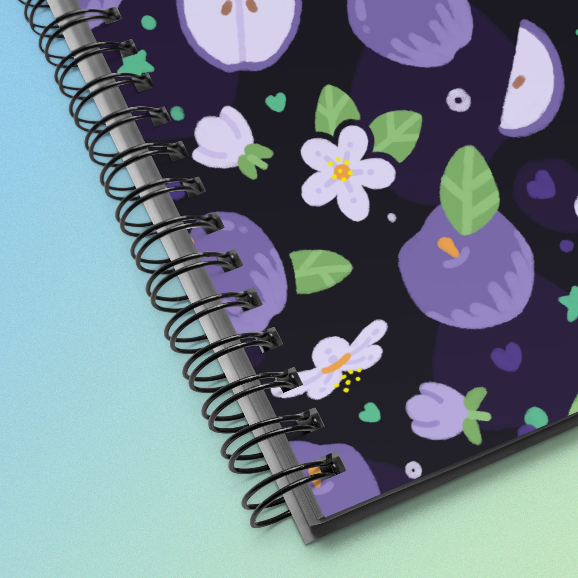 Close up of a spiral notebook with a colourful purple apples and apple blossoms pattern on a blue and green gradient background.