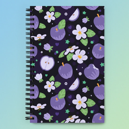 Spiral notebook with a colourful purple apples and apple blossoms pattern on a blue and green gradient background.