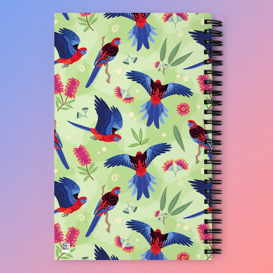 Spiral notebook with a colourful green, red, blue and pink Crimson Rosella pattern on a pink and blue gradient background.