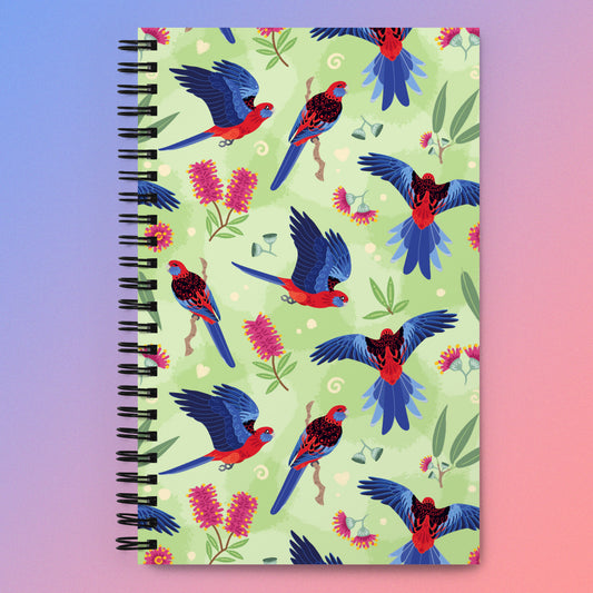 Spiral notebook with a colourful green, red, blue and pink Crimson Rosella pattern on a pink and blue gradient background.