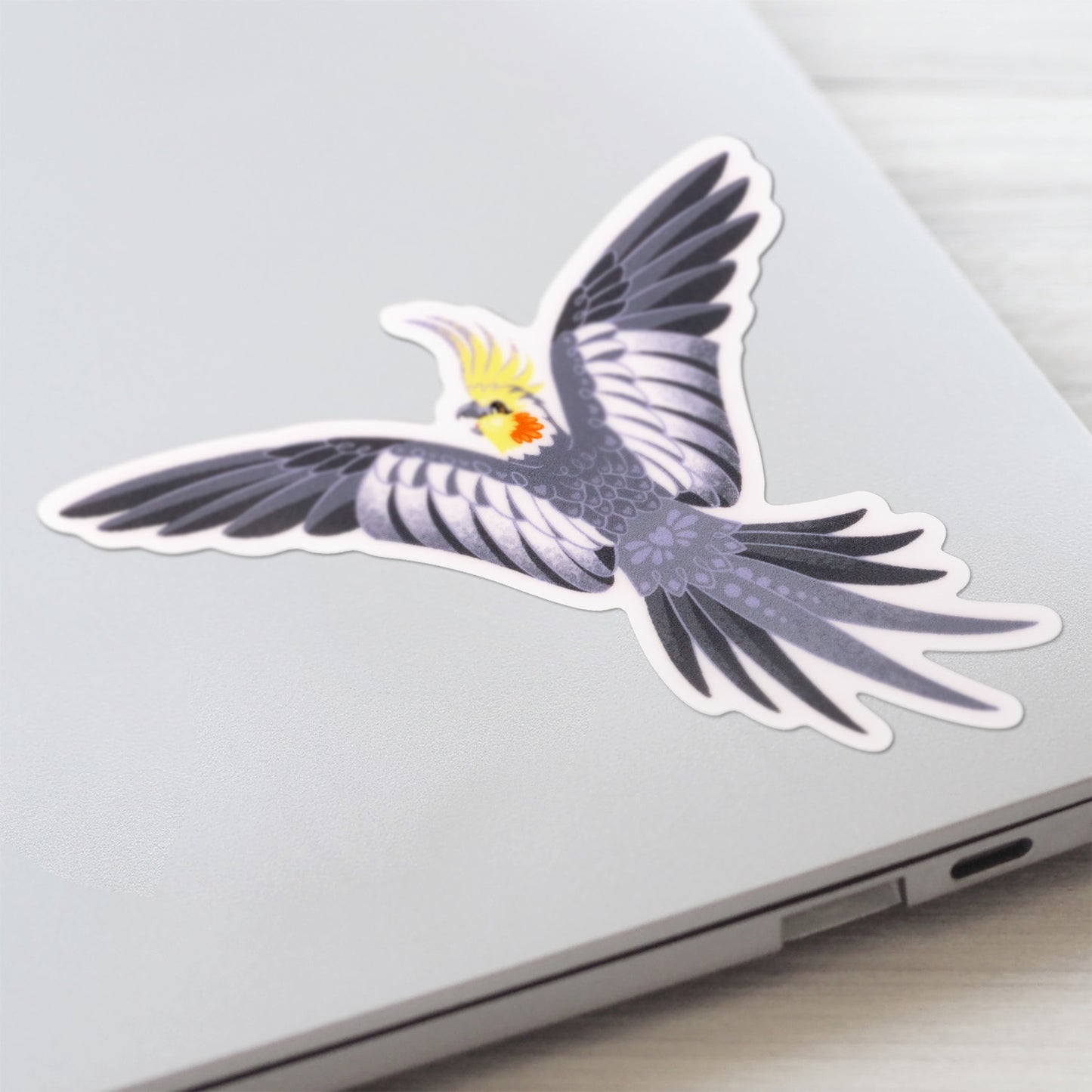 Close up of a colourful sticker of a male wild-type cockatiel spreading his wings wide stuck to a lid of a Macbook laptop on a textured white wood background.
