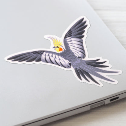 Close up of a colourful sticker of a male wild-type cockatiel spreading his wings wide stuck to a lid of a Macbook laptop on a textured white wood background.