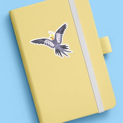 Colourful sticker of a male wild-type cockatiel spreading his wings wide stuck to a pale yellow journal on a blue background.