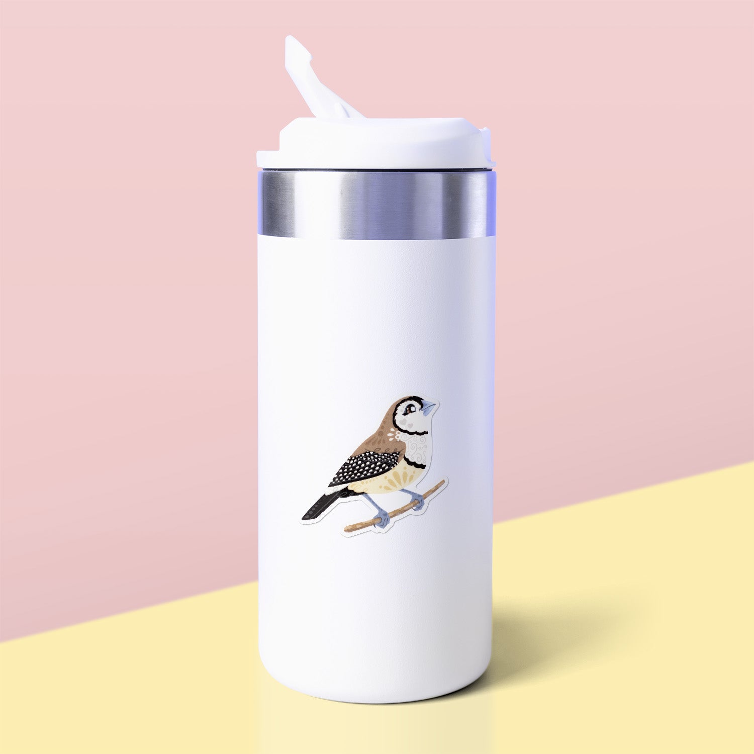 Colourful die-cut sticker of a double-barred owl finch stuck to a white metal drink bottle on a soft yellow table on a soft peach background.