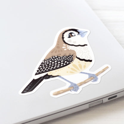 Colourful die-cut sticker of a double-barred owl finch stuck to a lid of a Macbook laptop on a textured white wood background.