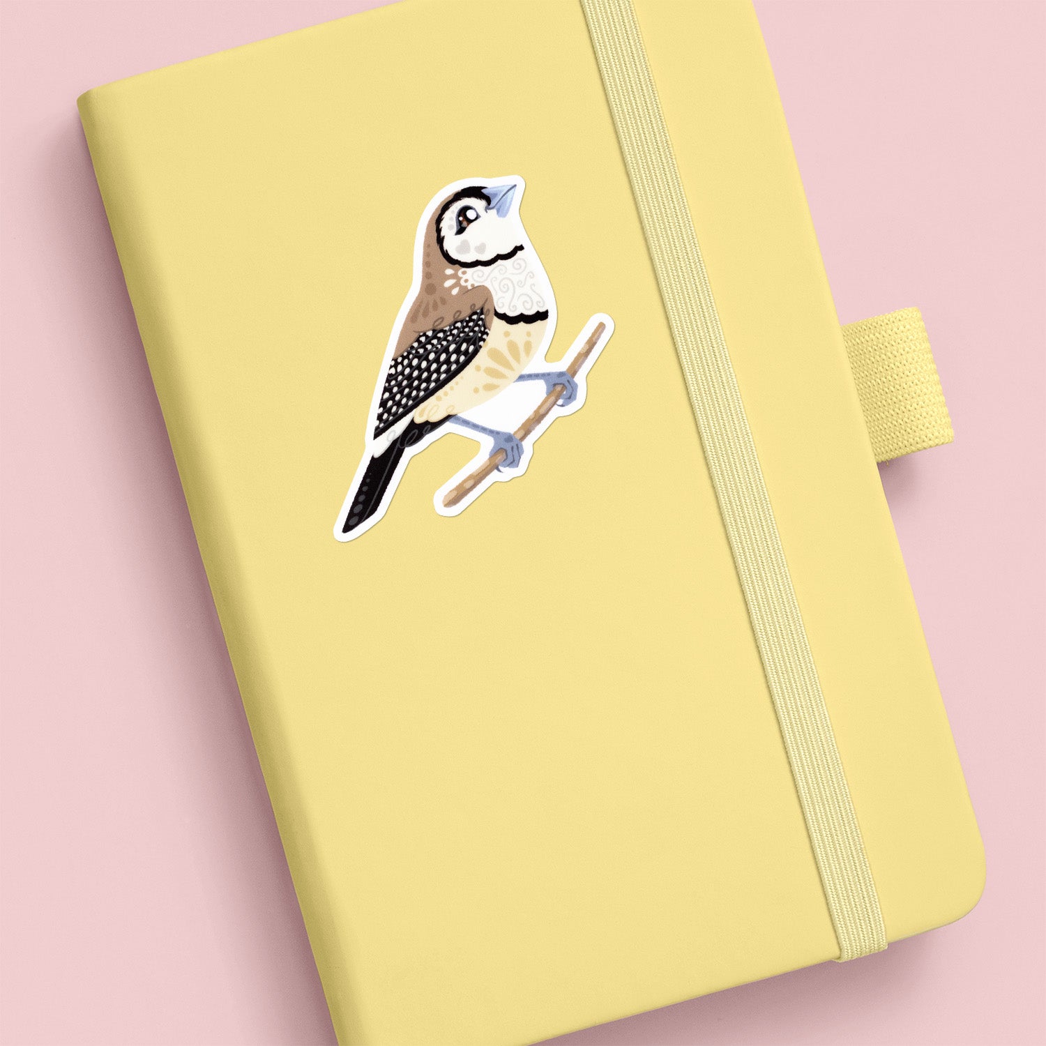 Colourful die-cut sticker of a double-barred owl finch stuck to a pale yellow journal on an soft peach background.