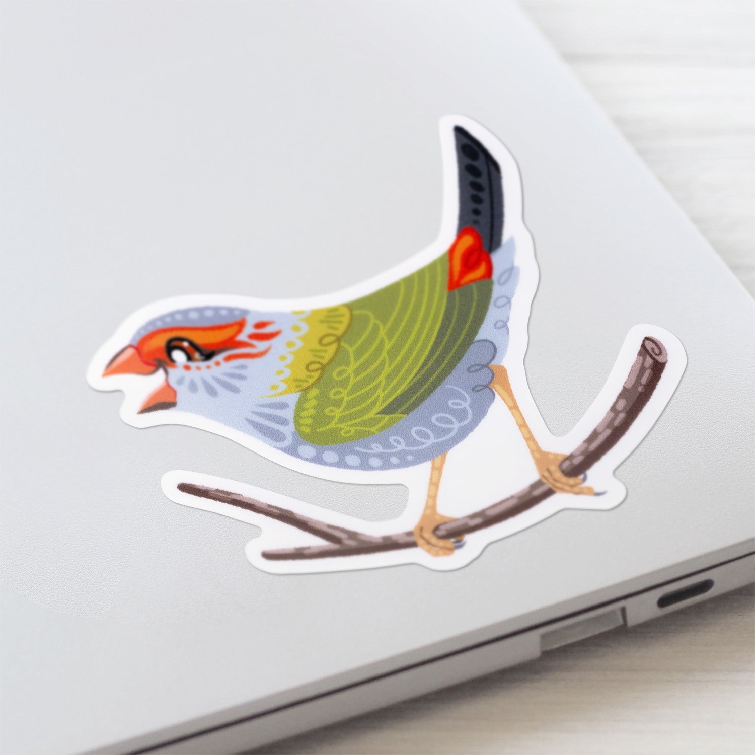 Colourful sticker of a red-browed firetail finch stuck to a lid of a Macbook laptop on a textured white wood background.
