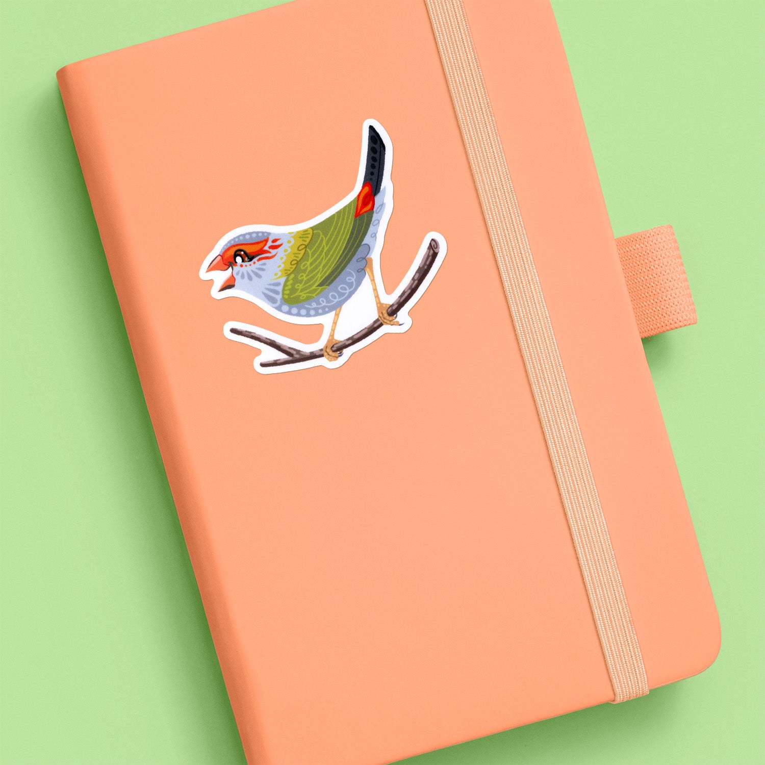 Colourful sticker of a red-browed firetail finch stuck to a pale orange journal on aa soft green background.