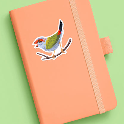 Colourful sticker of a red-browed firetail finch stuck to a pale orange journal on aa soft green background.