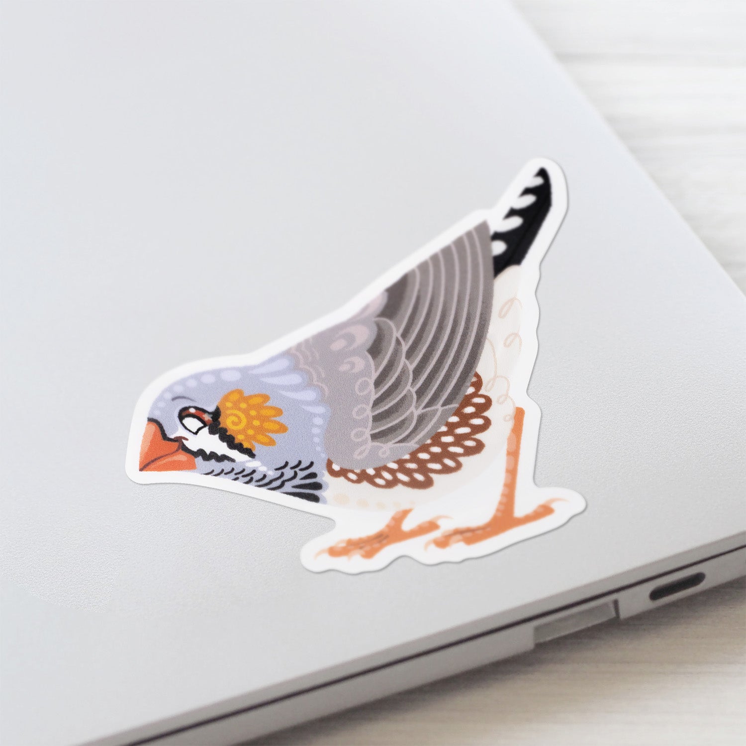 Closeup of a colourful die-cut sticker of a male Australian zebra finch stuck to a lid of a Macbook laptop on a textured white wood background.