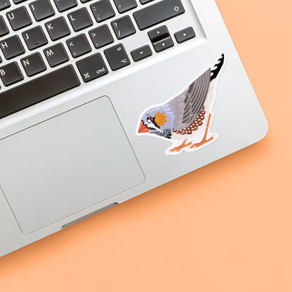 Colourful die-cut sticker of a male Australian zebra finch stuck below the keyboard of a Macbook laptop on a yellow background.