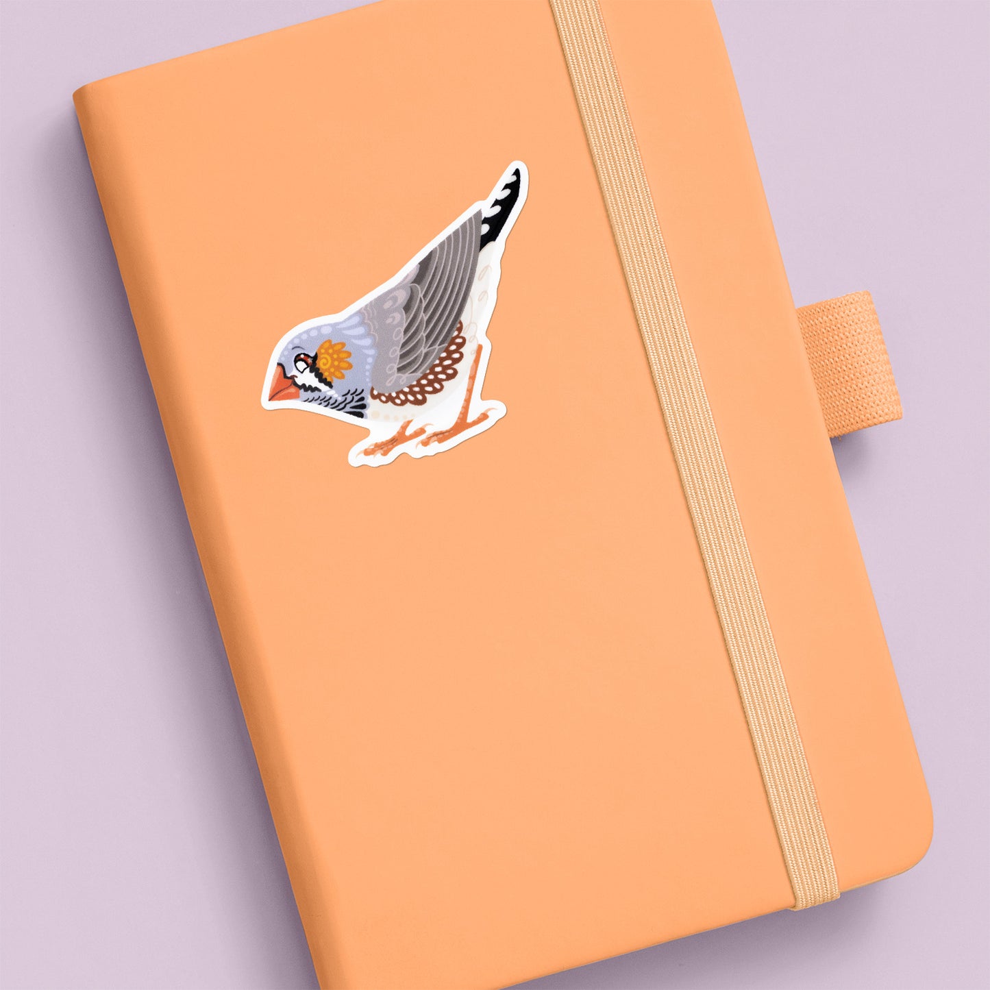 Colourful die-cut sticker of a male Australian zebra finch stuck to a pale orange journal on an soft lilac background.