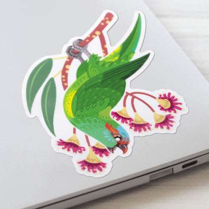 Closeup of a colourful sticker of a musk lorikeet perched amongst pink eucalyptus flowers stuck to a lid of a Macbook laptop on a textured white wood background.