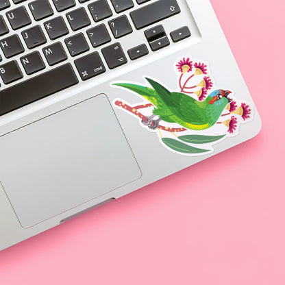 Colourful sticker of a musk lorikeet perched amongst pink eucalyptus flowers stuck below the keyboard of a Macbook laptop on a pink background.