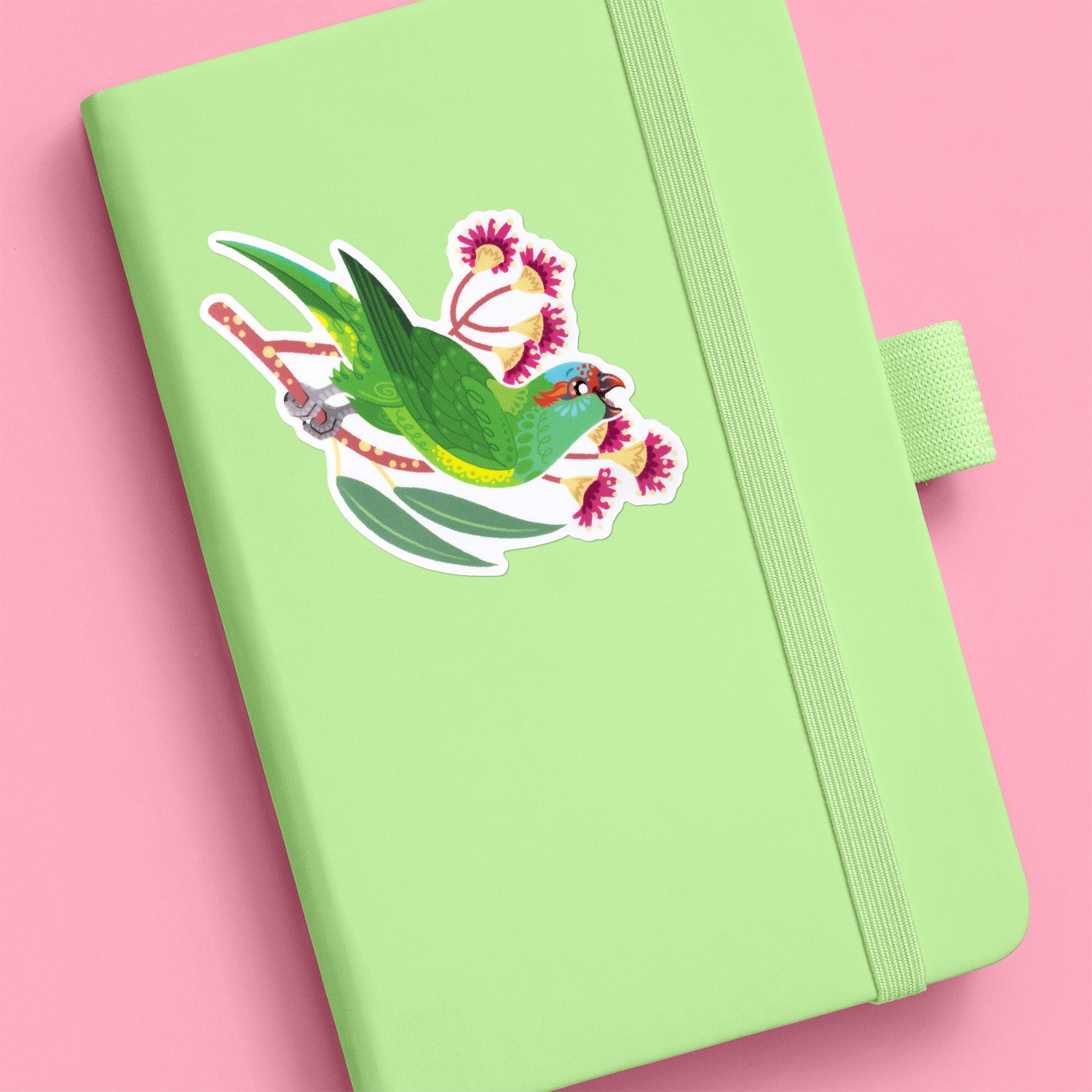 Colourful sticker of a musk lorikeet perched amongst pink eucalyptus flowers stuck to a pale green journal on a pink background.