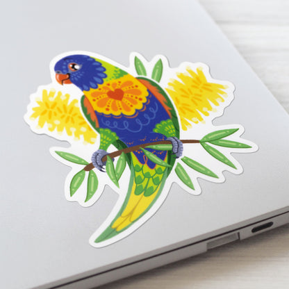 Closeup of a colourful sticker of a rainbow lorikeet with yellow bottlebrush flowers stuck to a lid of a Macbook laptop on a textured white wood background.