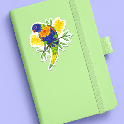 Colourful sticker of a rainbow lorikeet with yellow bottlebrush flowers stuck to a pale green journal on a lilac background.