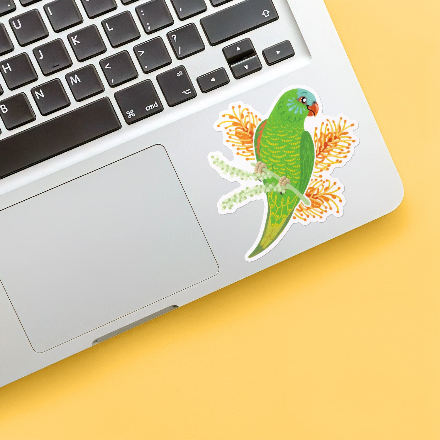 A colourful sticker of a scaly-breasted lorikeet with orange grevillea flowers stuck below the keyboard of a Macbook laptop on a yellow background.