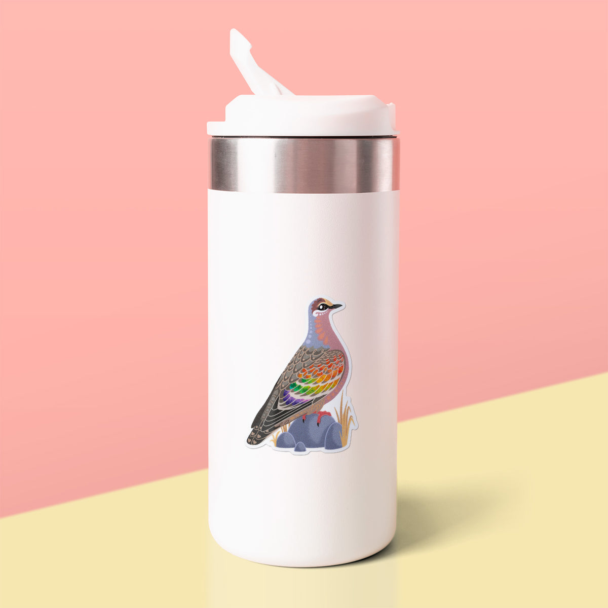 Colourful die-cut sticker of a male common bronzewing pigeon stuck to a white metal drink bottle on a soft yellow table on a peach background.