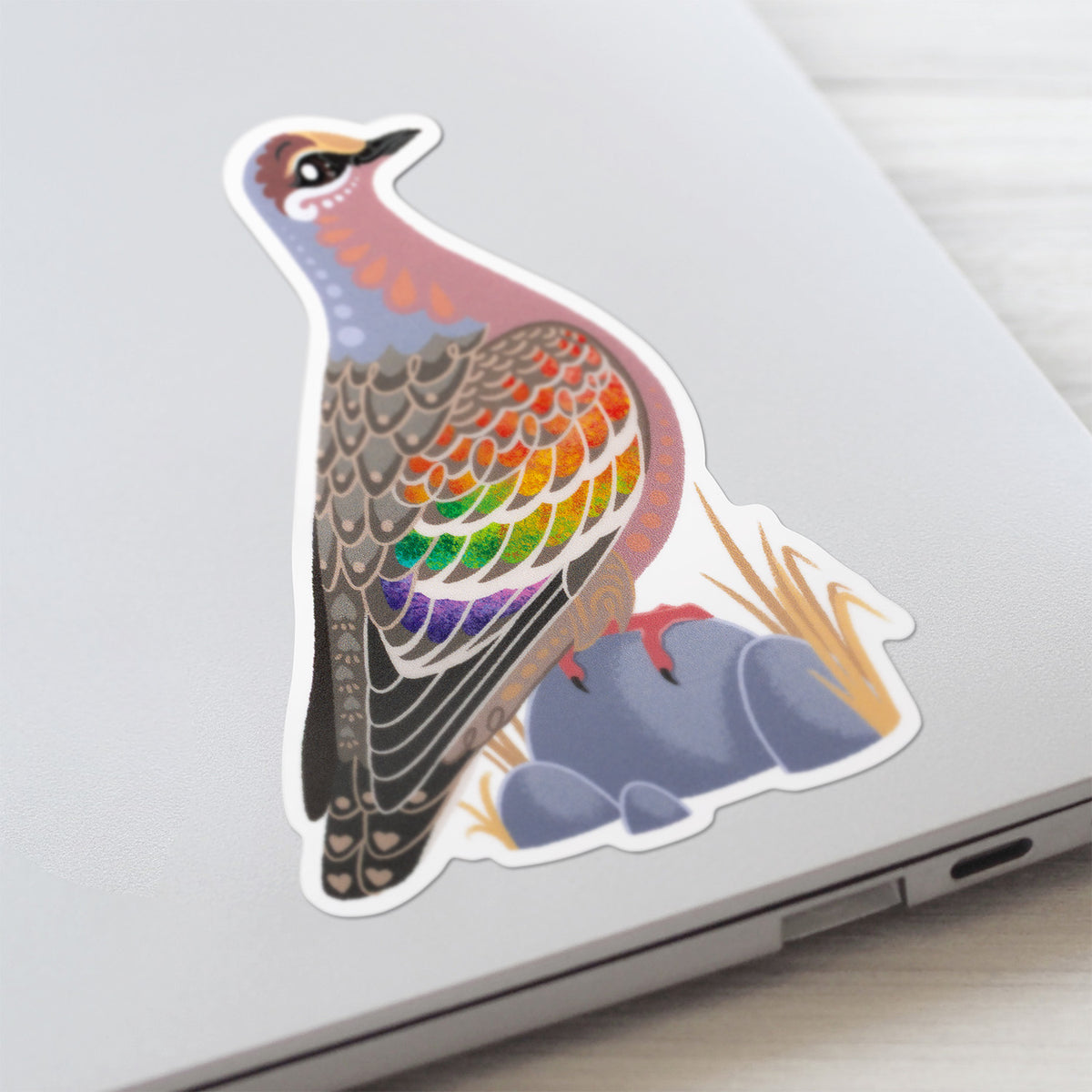 Colourful die-cut sticker of a male common bronzewing pigeon stuck to a lid of a Macbook laptop on a textured white wood background.